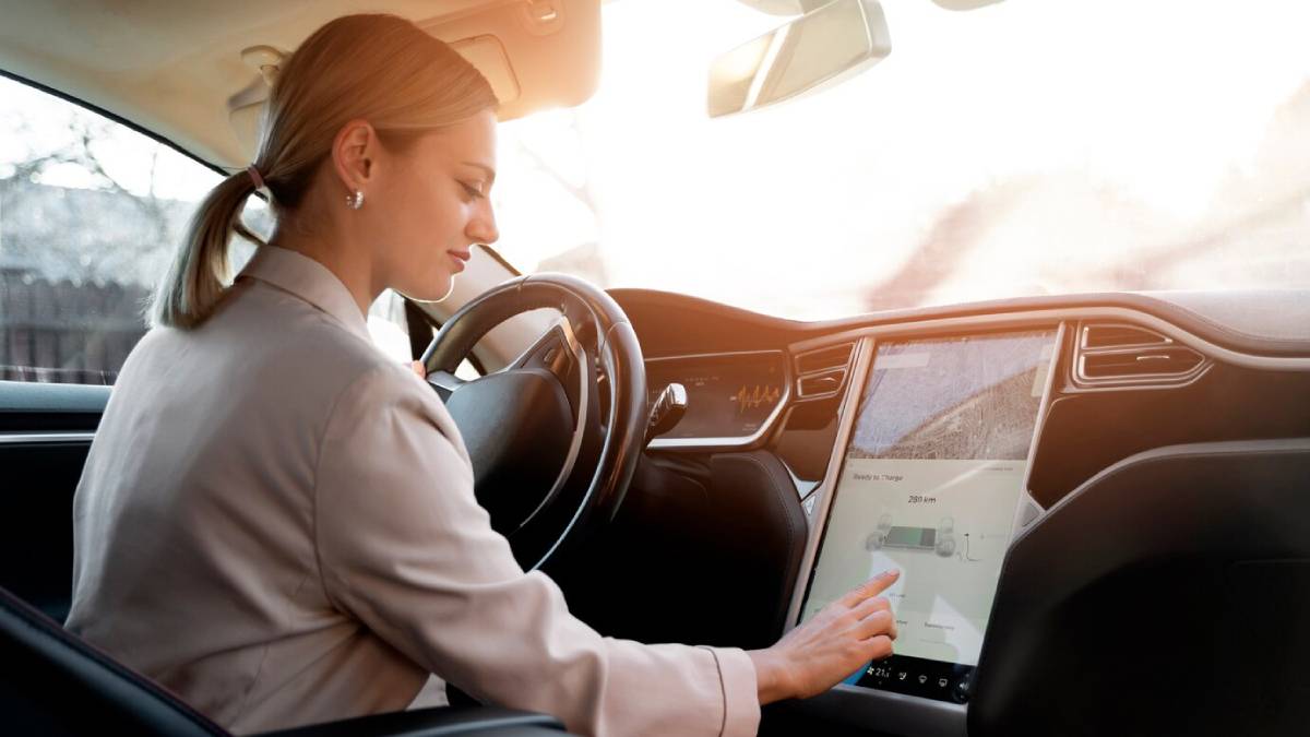 A woman in a beige jacket sits in a car, interacting with a large touchscreen displaying navigation details. Sunlight streams through the window.