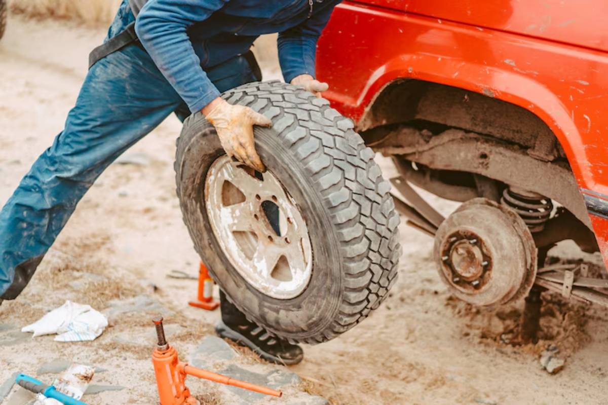 A person in blue overalls lifts a tire to install it on a red car, surrounded by tools on a sandy road.