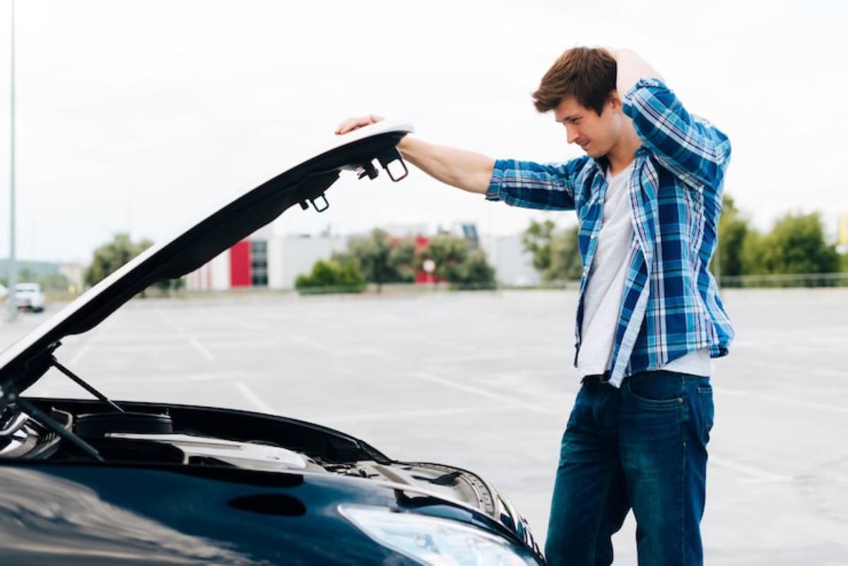 A person in a plaid shirt stands beside a car with its hood open, looking puzzled in an empty parking lot.
