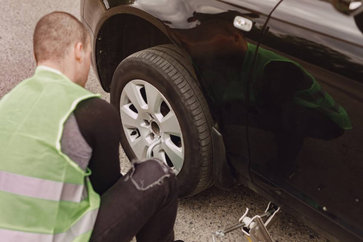 A person wearing a reflective vest crouches near a black car, inspecting the tire and preparing to change it on the roadside.