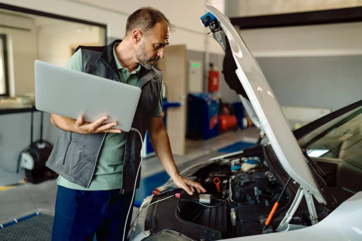 A mechanic examines the engine of a car while holding a laptop, engaged in troubleshooting within a garage setting.
