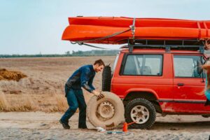 A person replaces a tire on a red SUV parked on a dirt road, with orange kayaks secured on the roof and open fields in the background.