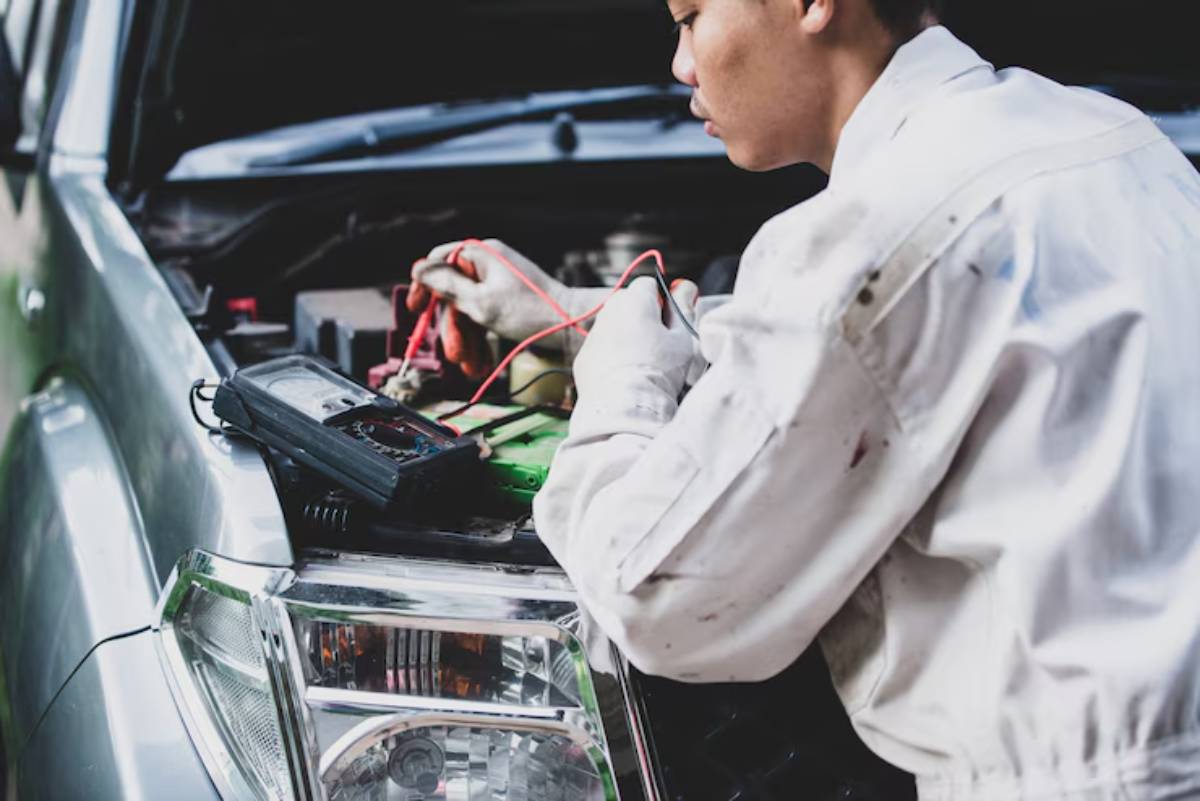 A mechanic in a white suit checks a car battery with red and black wires, using a multimeter on the hood of a silver vehicle.