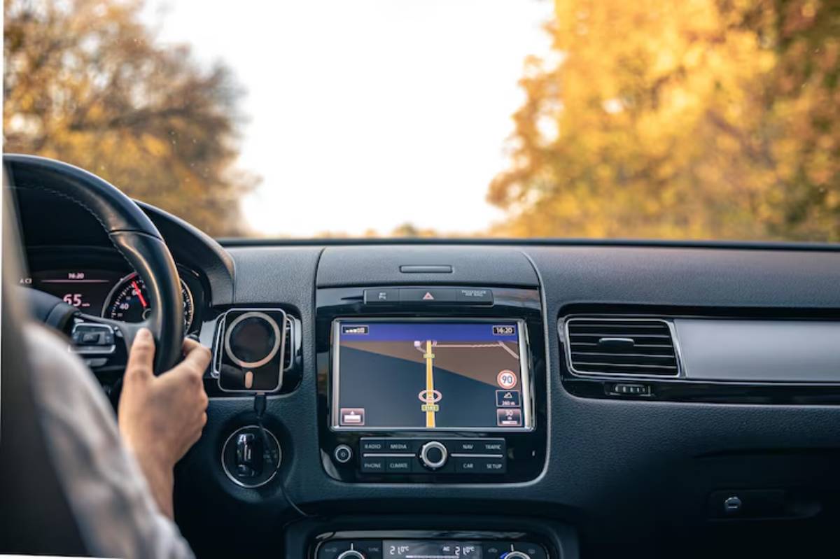 A driver's hands on a steering wheel, with a GPS screen displaying navigation directions and autumn scenery outside the windshield.