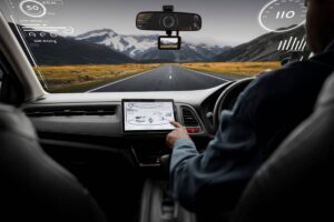 A person interacts with a touchscreen in a self-driving car, with a scenic mountain road visible through the windshield.