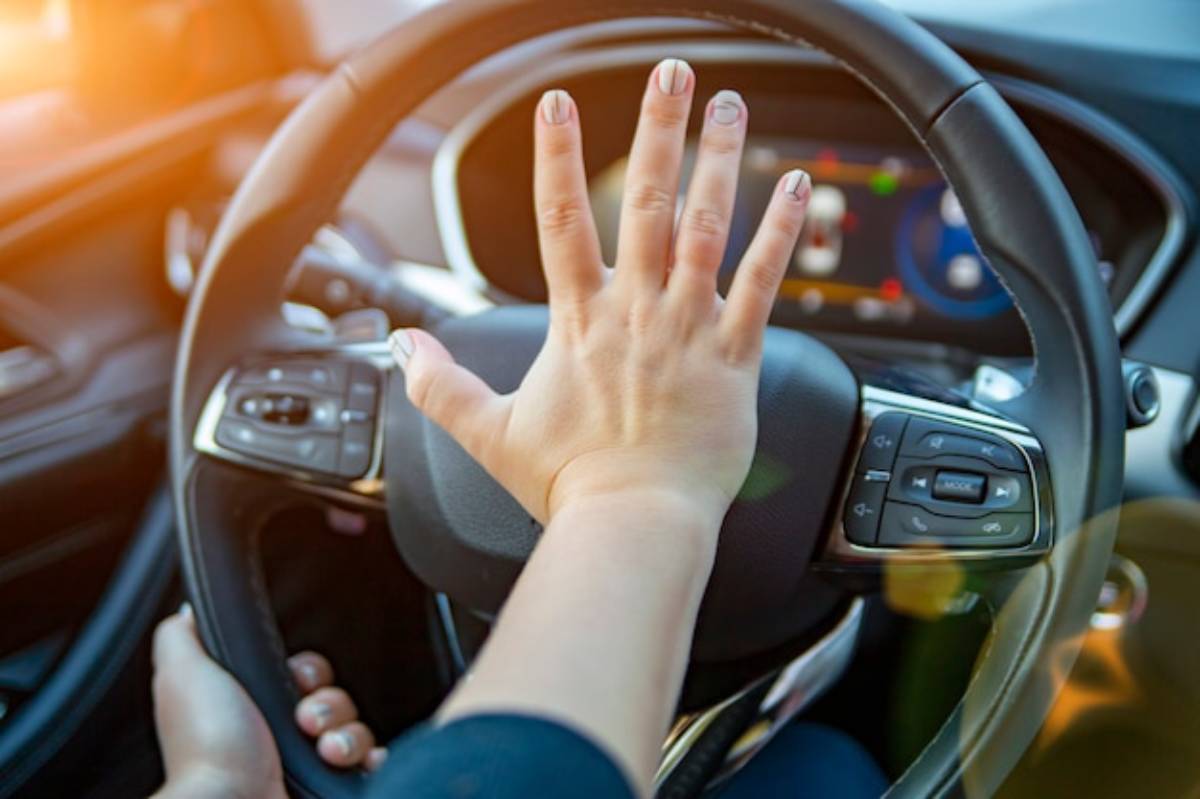 A hand signaling 'stop' on a car steering wheel, with a colorful dashboard display and sunlight filtering in through the window.