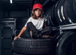 A child in a red cap sits cross-legged atop a stack of tires, surrounded by more tires in an industrial garage setting.