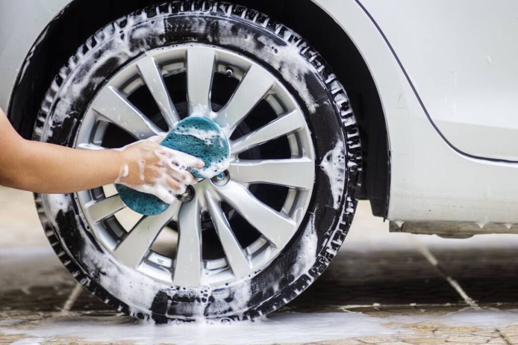 A person uses a sponge to clean a car tire, covered in soap suds, while water accumulates on the ground beneath.