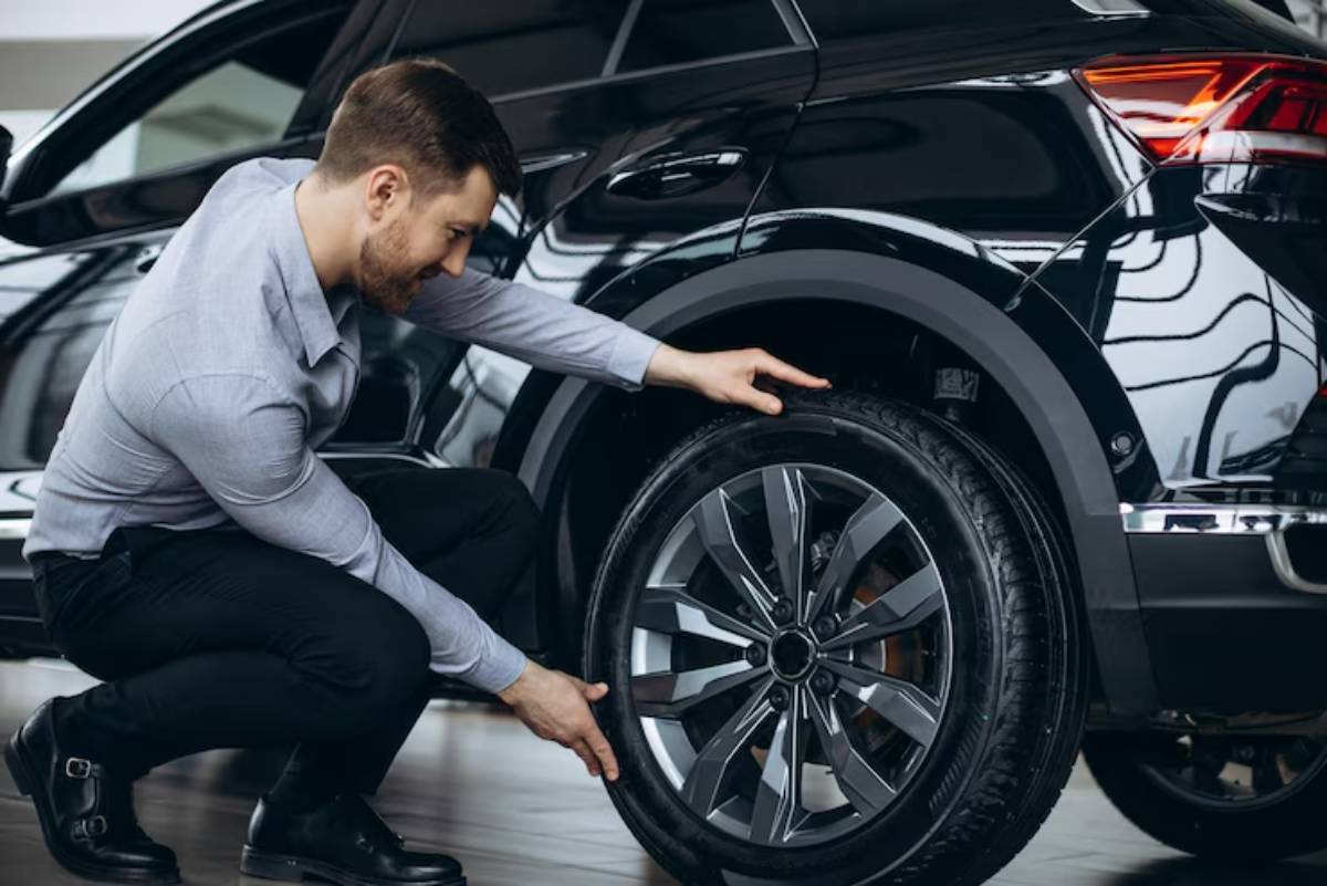 A man in a light shirt inspects the tire of a sleek black car, kneeling in a modern, well-lit garage environment.