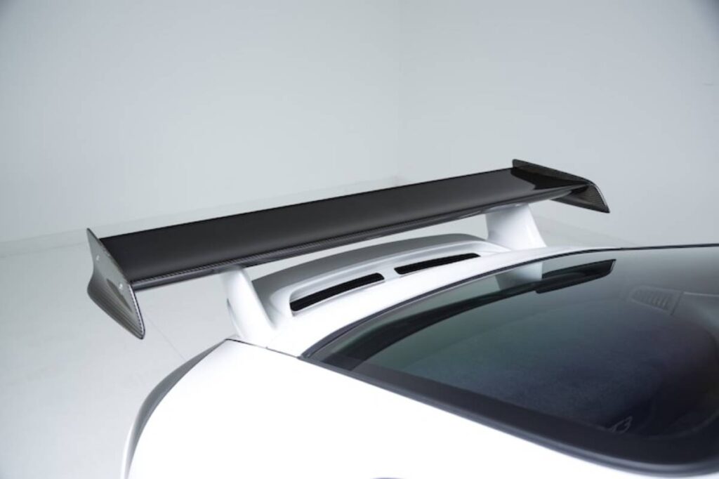A close-up of a sleek white sports car rear featuring a prominent black carbon fiber wing on top of the rear deck.