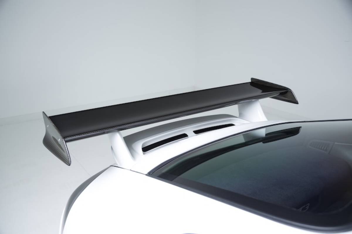 A close-up of a sleek white sports car rear featuring a prominent black carbon fiber wing on top of the rear deck.