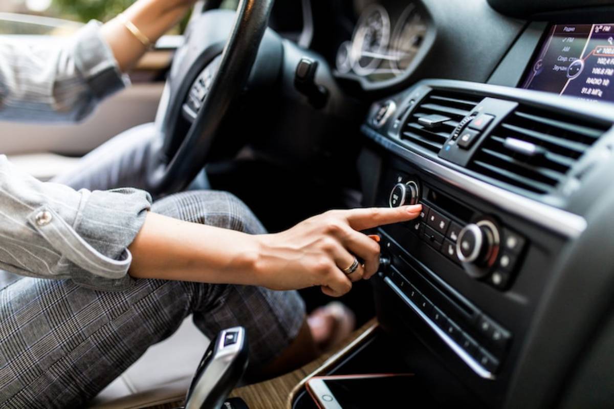 A person in a car adjusts the radio, with hands on the steering wheel and a smartphone on the console.