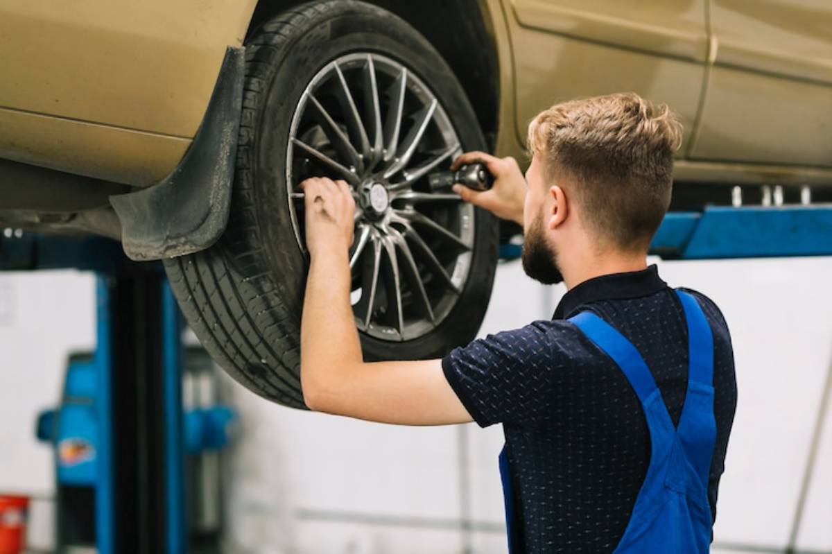 A mechanic in blue overalls works on a raised car, using a wrench to tighten the wheel. The vehicle is light brown, showcasing its alloy rims.