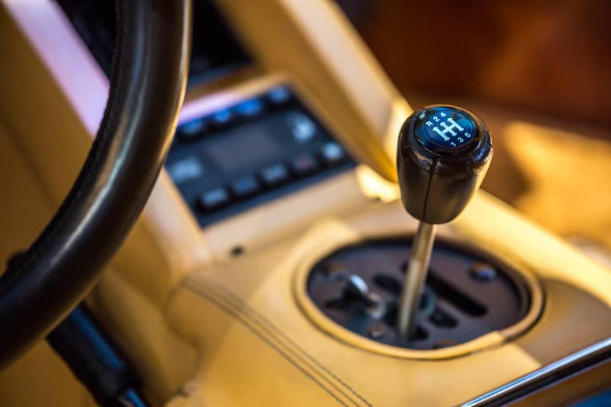 Close-up of a manual gear stick in a car interior, featuring a black shift knob with gear markings and a leather-wrapped steering wheel.