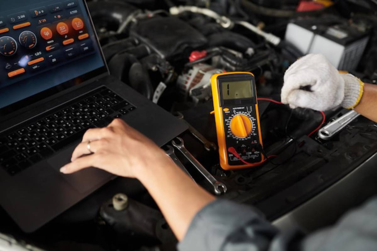 A mechanic tests electrical components using a multimeter, with a laptop displaying diagnostic information, in a car's engine bay.