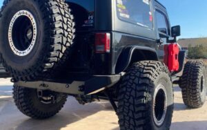 A modified black Jeep with oversized tires, a red fuel canister, and various off-road stickers, parked outdoors.