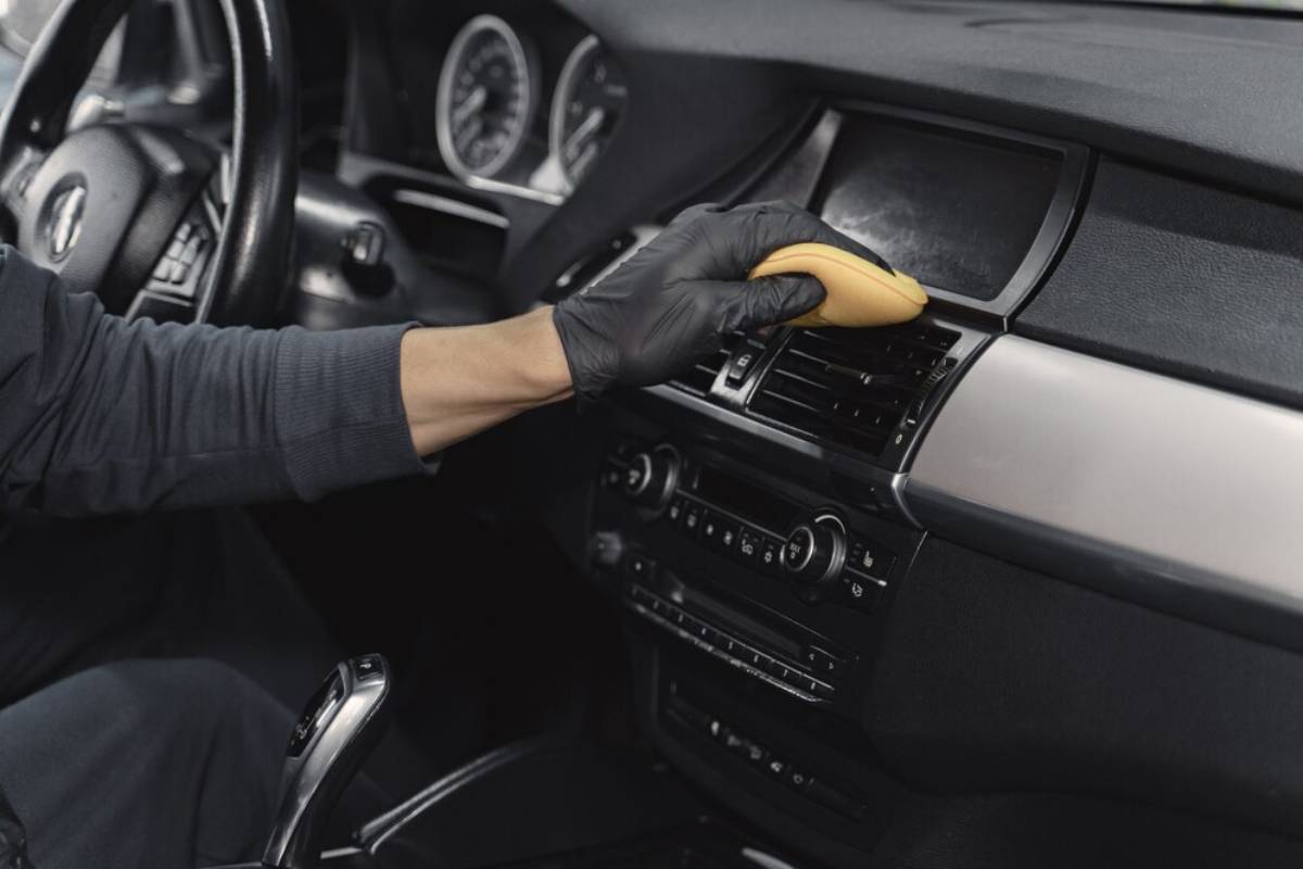 A person in a black glove cleans the dashboard and air vents of a car's interior using a yellow cleaning sponge.