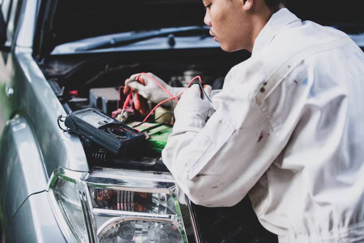 A mechanic in a white coverall works on a car battery, using a multimeter and connecting cables. The car's hood is open.