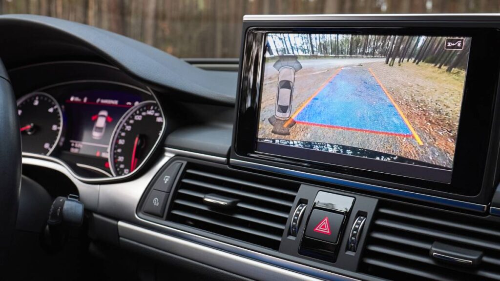 Interior view of a car displaying a rearview camera screen with guidelines on a forested road, showing safe maneuvering.