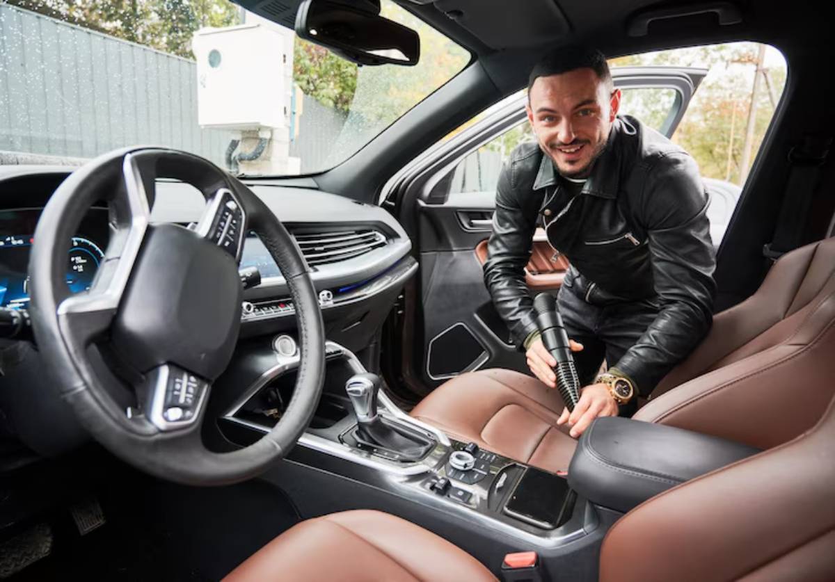 A person in a black leather jacket is using a vacuum to clean the brown leather interior of a car.