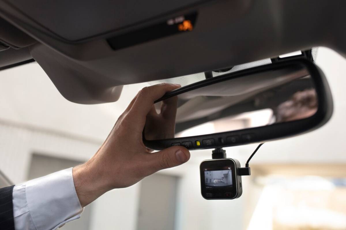 A hand adjusts a car's rearview mirror, which features a small digital camera attached beneath it.