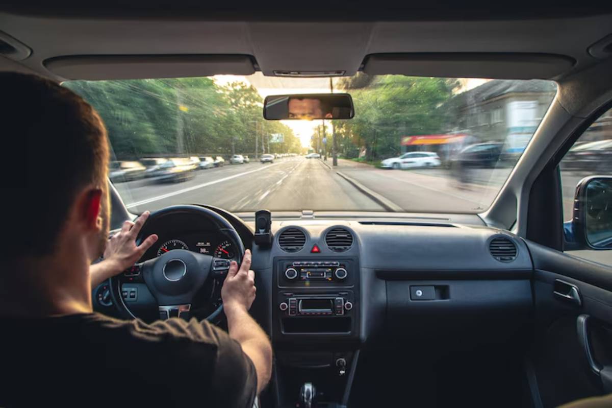 A driver with short hair navigates a city street, hands on the wheel, as sunlight filters through trees lining the road.