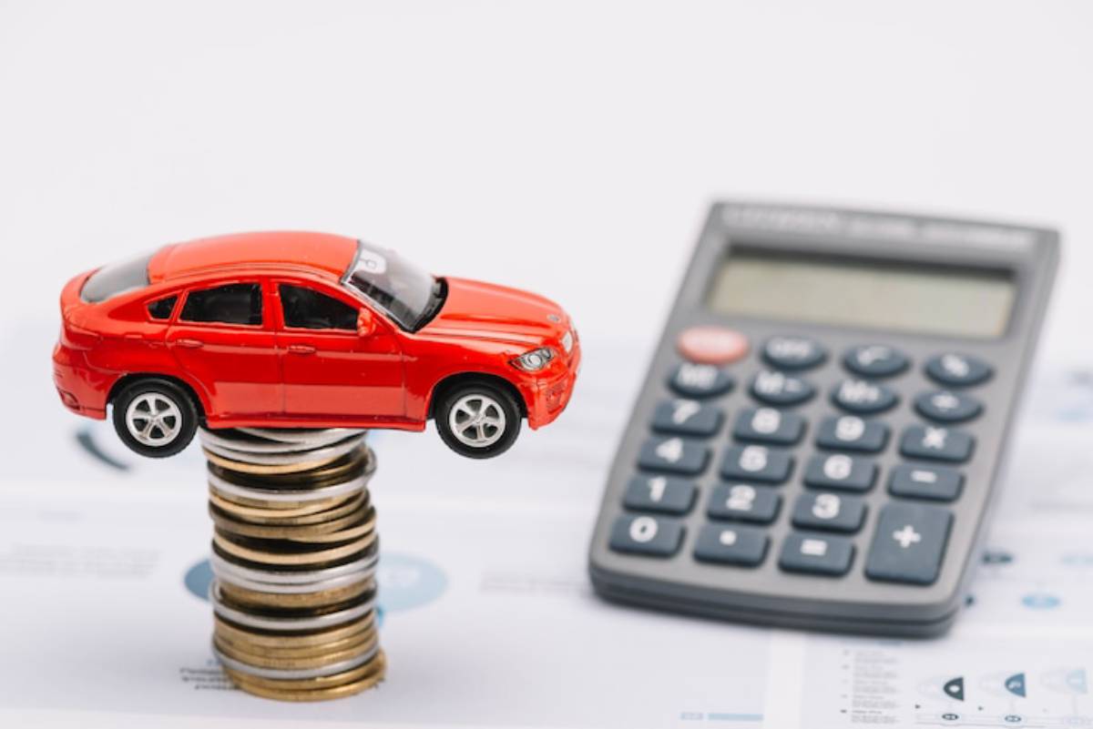 A toy red car balanced on a stack of coins, with a calculator nearby, symbolizing car finance or budgeting concepts.