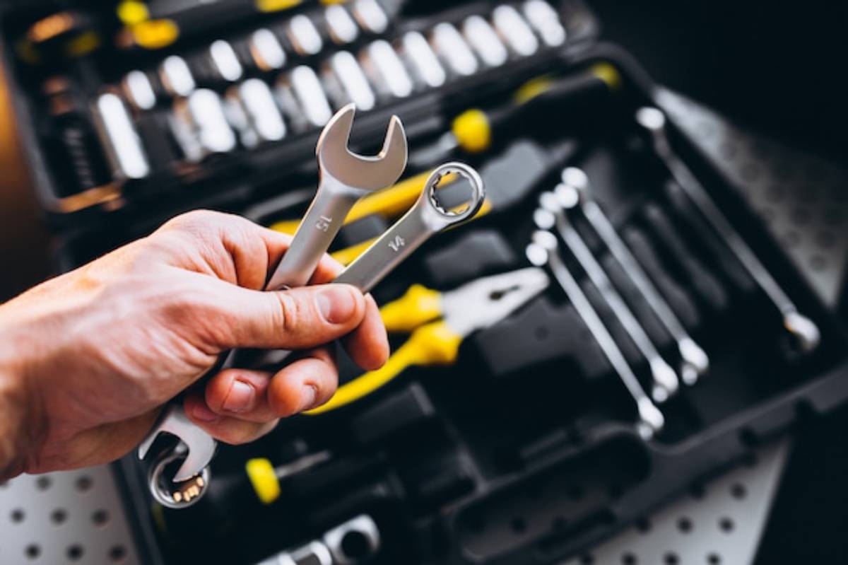 A hand holds two metallic wrenches above a tool kit filled with various tools and sockets, highlighting a DIY or repair setting.