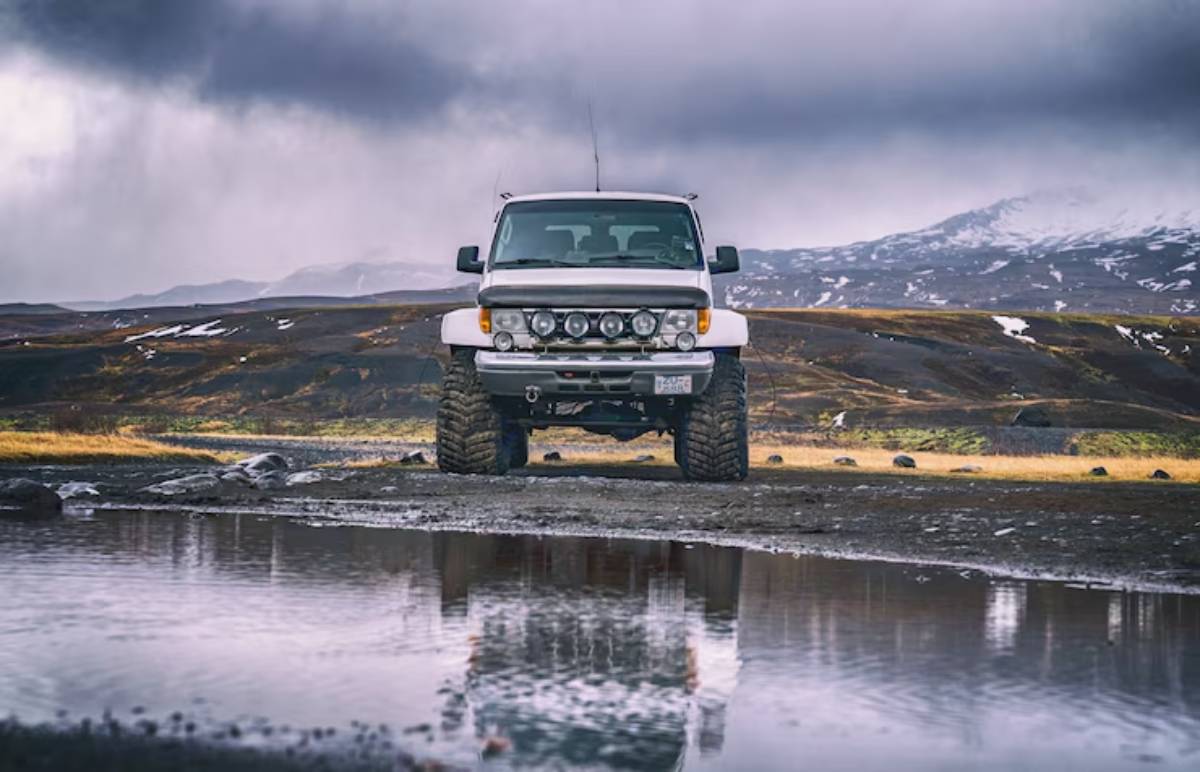 A rugged white off-road vehicle parked near a reflective puddle, with mountainous terrain and dramatic clouds in the background.