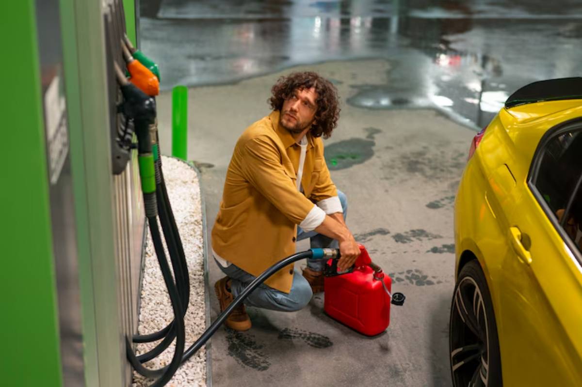 A person in a tan jacket fills a red gasoline canister at a fuel station next to a yellow car, with wet pavement in the background.