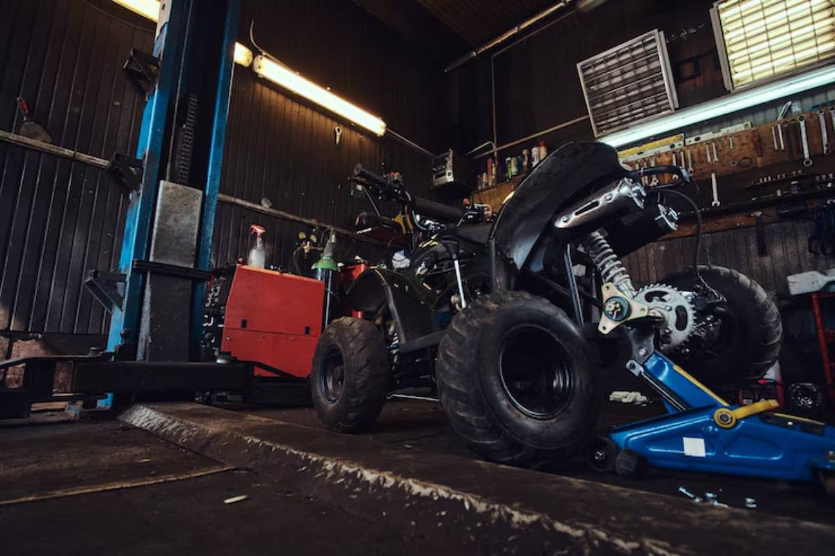A black quad bike on a workshop floor, surrounded by tools and equipment, with a blue lift and red toolbox in a dimly lit garage.