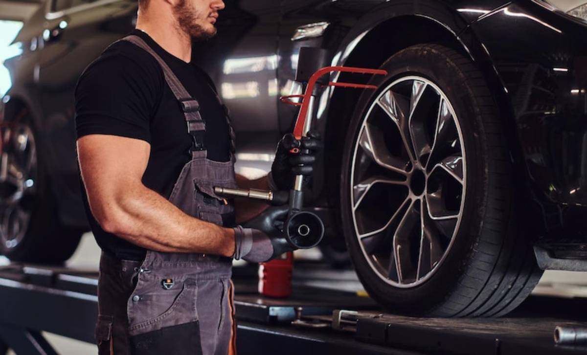 A mechanic in gray overalls holds a tool and inspects a car's tire on a workshop lift, with a sleek black vehicle in the background.
