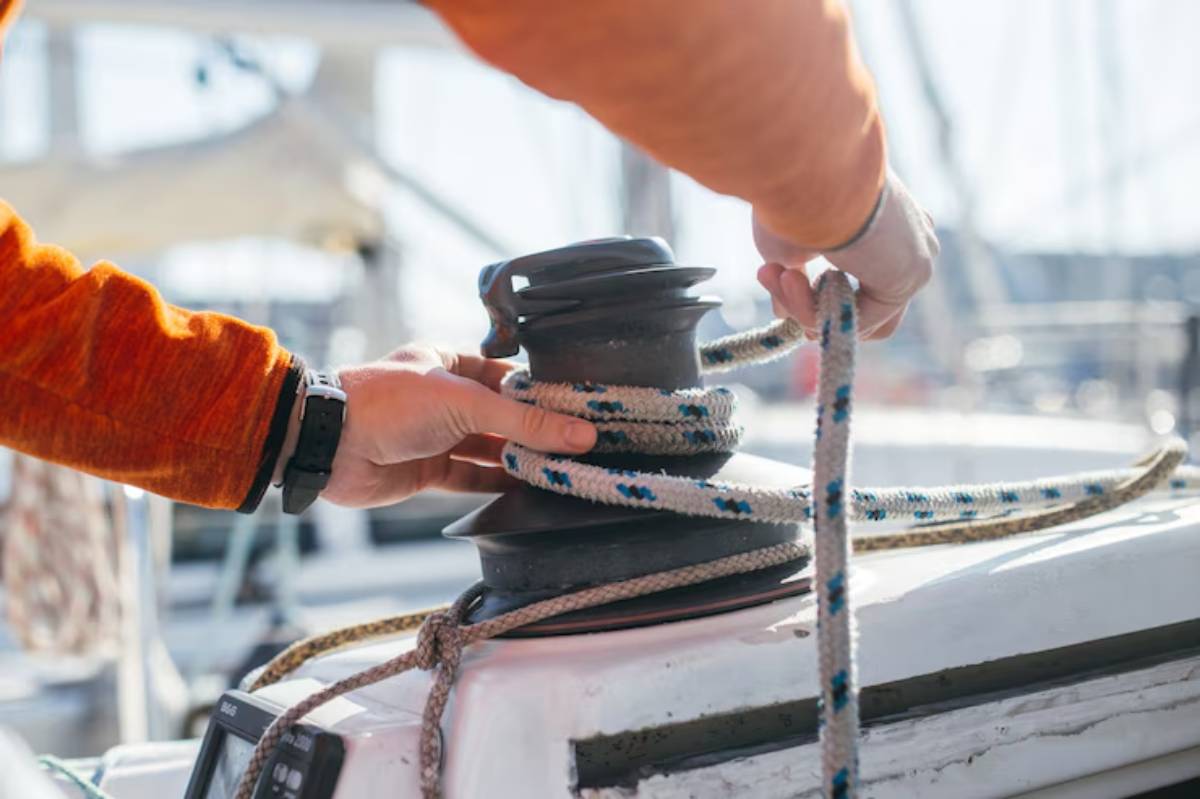 A person in an orange shirt is securing a rope around a boat winch on a sunny day at the marina.