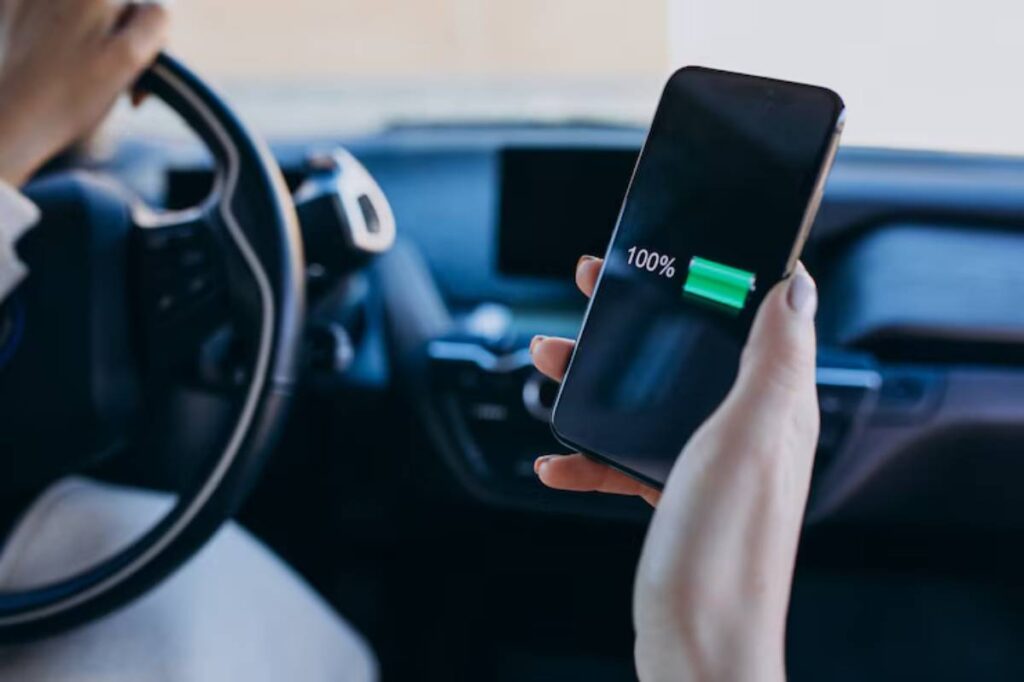 A hand holding a smartphone displaying a full battery (100%) while driving a car, with the steering wheel partially visible.
