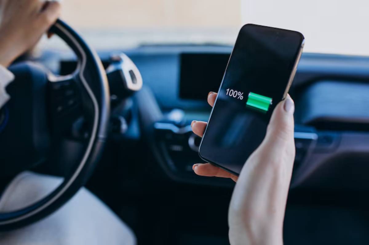 A hand holding a smartphone displaying a full battery (100%) while driving a car, with the steering wheel partially visible.