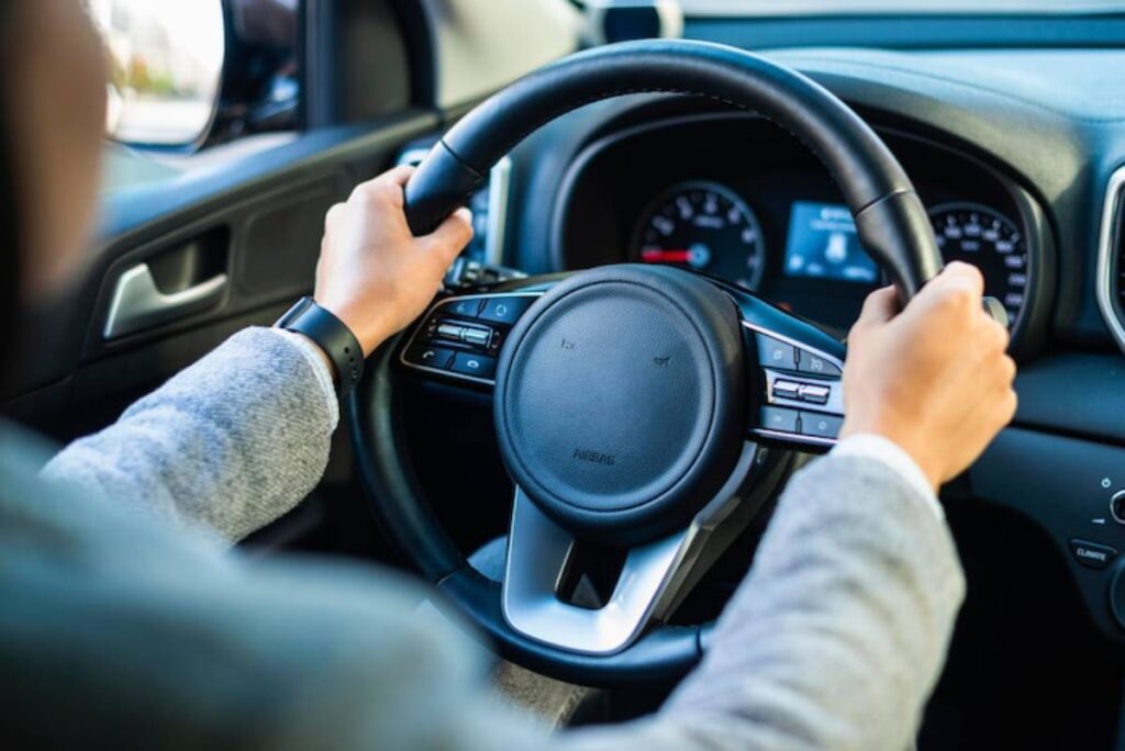 A driver's hands grasp the steering wheel of a modern car, with a dashboard displaying speed and controls visible in the background.