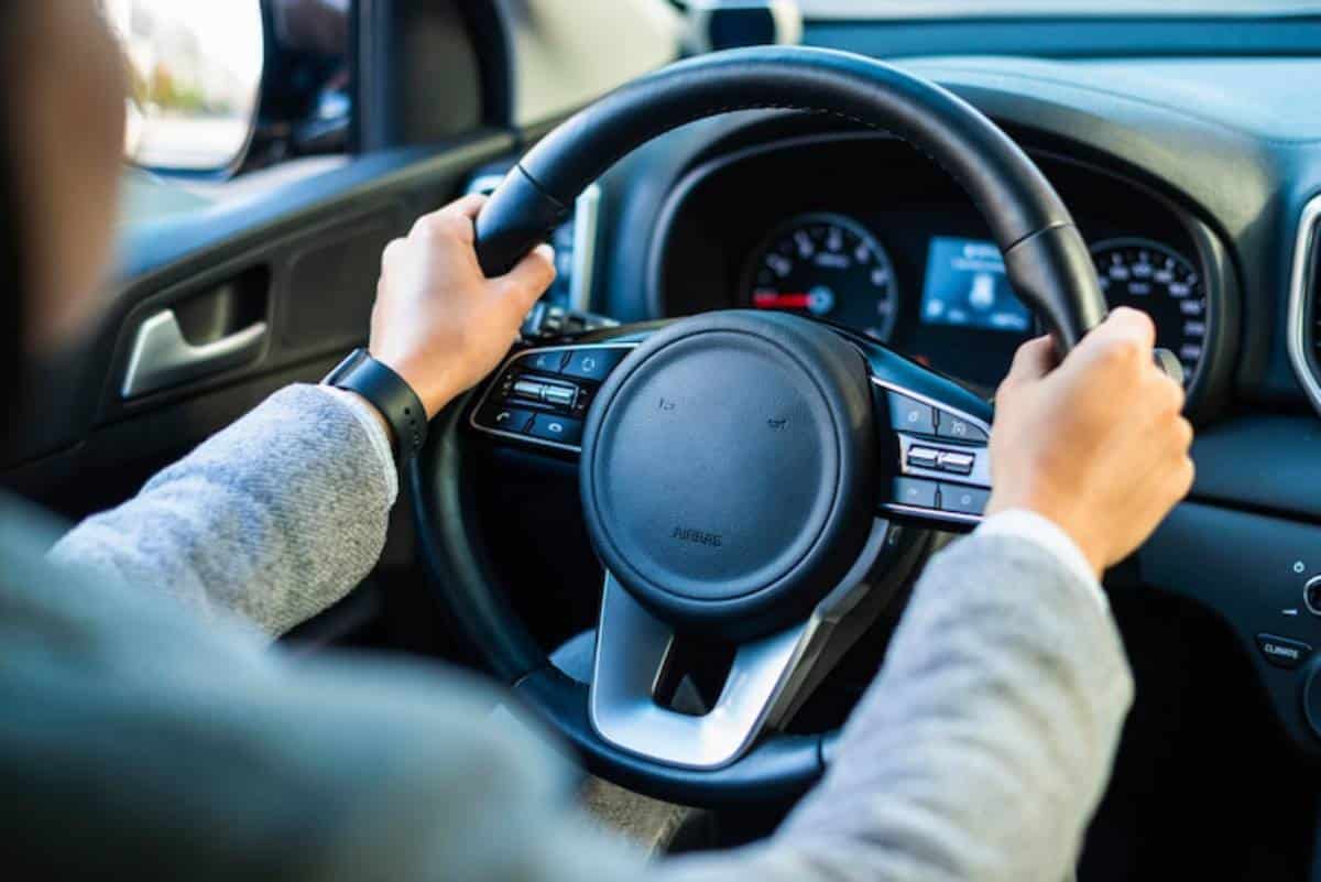 A driver's hands grasp the steering wheel of a modern car, with a dashboard displaying speed and controls visible in the background.