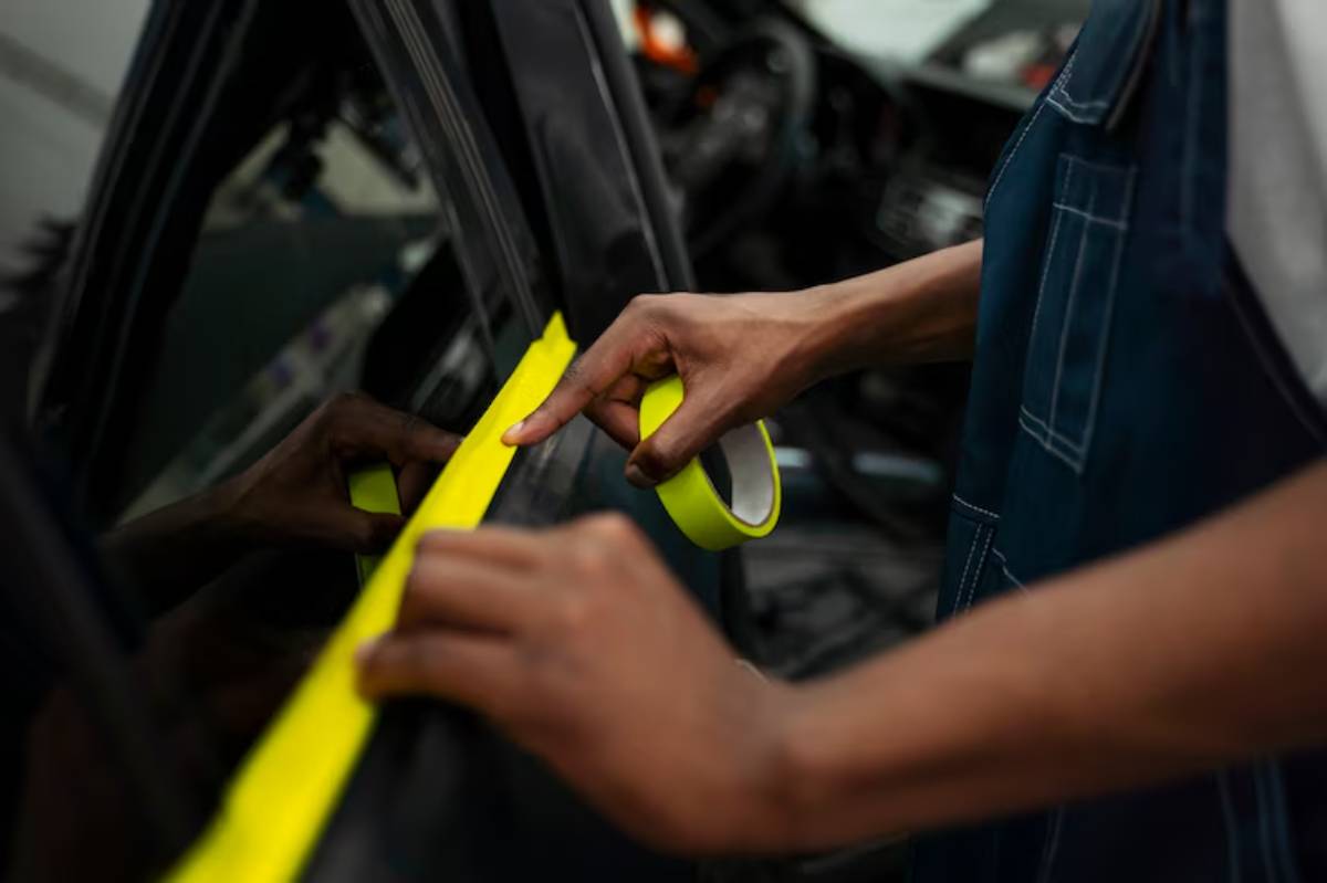 A person applies bright yellow tape along the edge of a car window, preparing for painting or detailing work inside a vehicle.