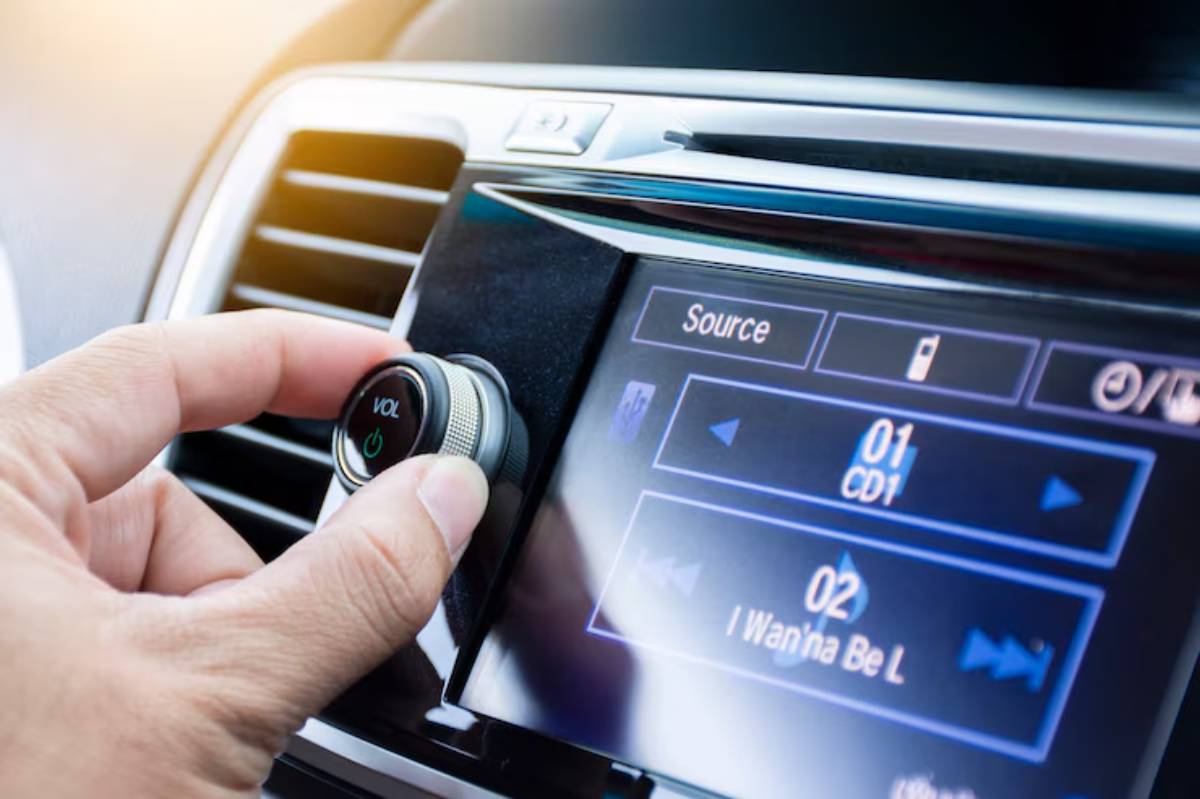 A hand adjusting the volume knob on a car audio system, with a digital display showing track information and options.