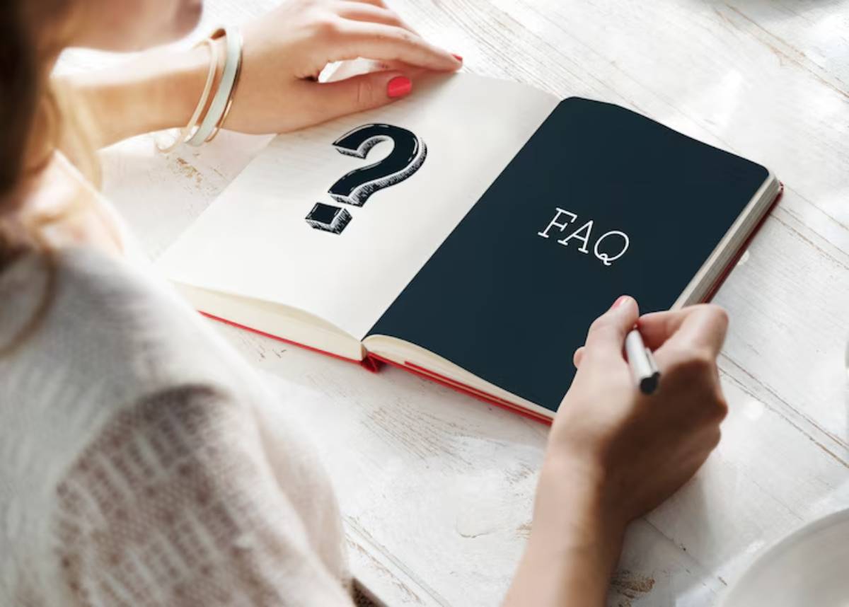 A person writing in a notebook labeled FAQ, alongside a large question mark, on a wooden table.