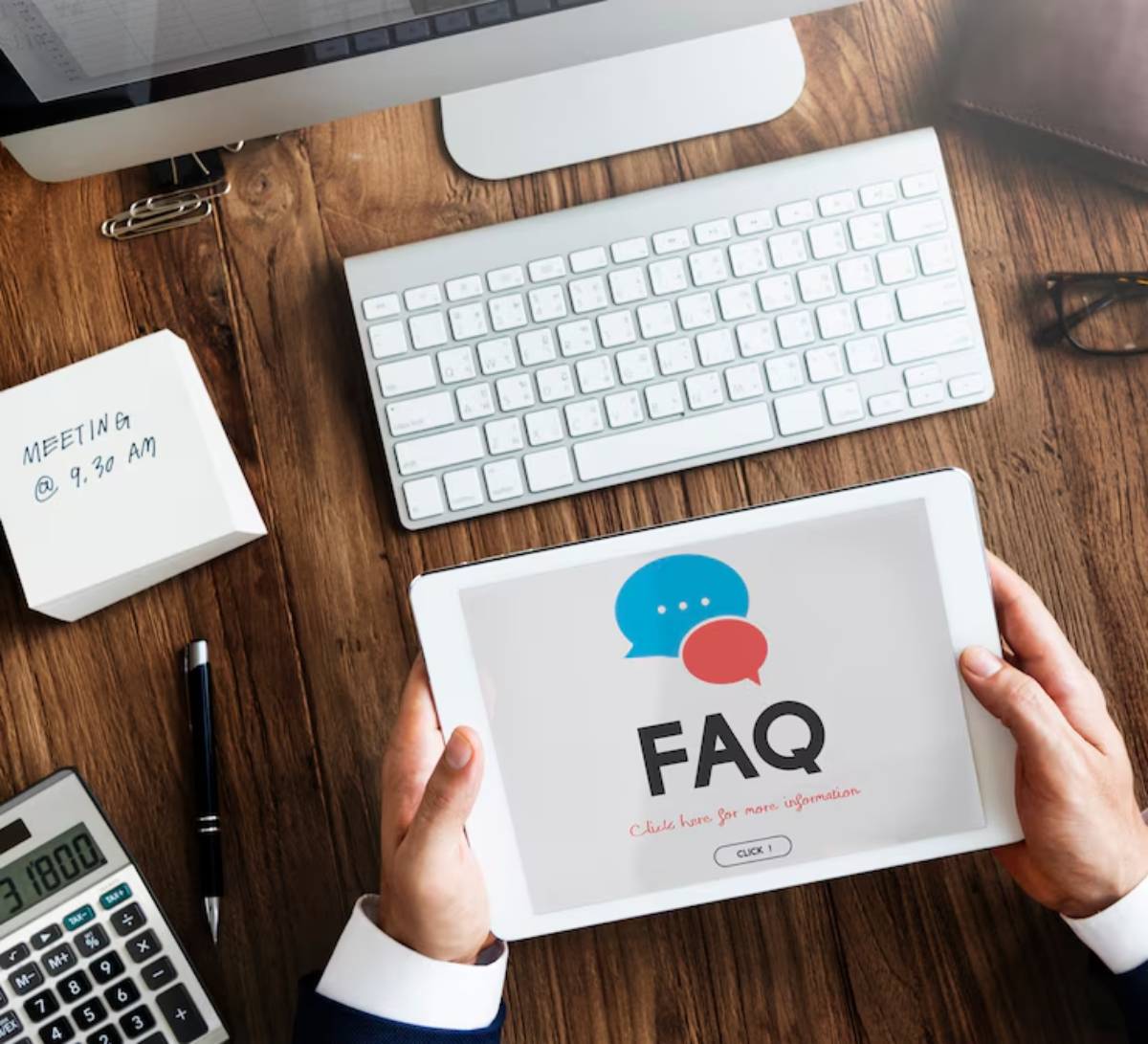 A person holds a tablet displaying FAQ with speech bubbles, sitting at a desk with a keyboard, planner, and calculator.