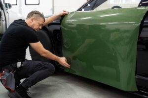 A technician applies a glossy green vinyl wrap to the side of a vehicle in a modern automotive workshop.