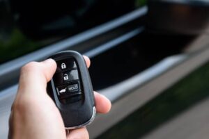 A hand holds a car key fob with lock, unlock, and hold buttons, positioned near a vehicle's door.