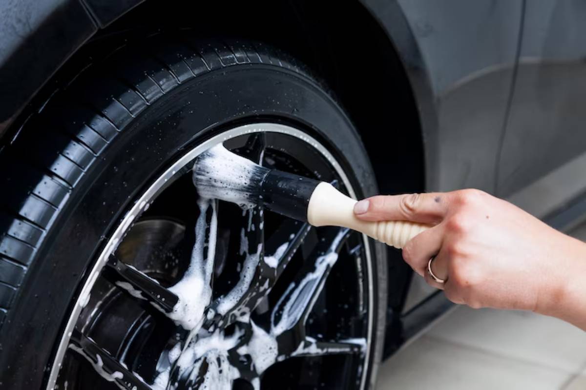 A hand holding a brush cleans a black car tire covered in soapy foam, highlighting a shiny rim and polished surface.