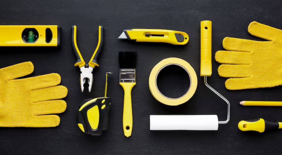A flat lay of yellow-handled tools including gloves, pliers, a level, a paintbrush, and a tape measure on a dark background.