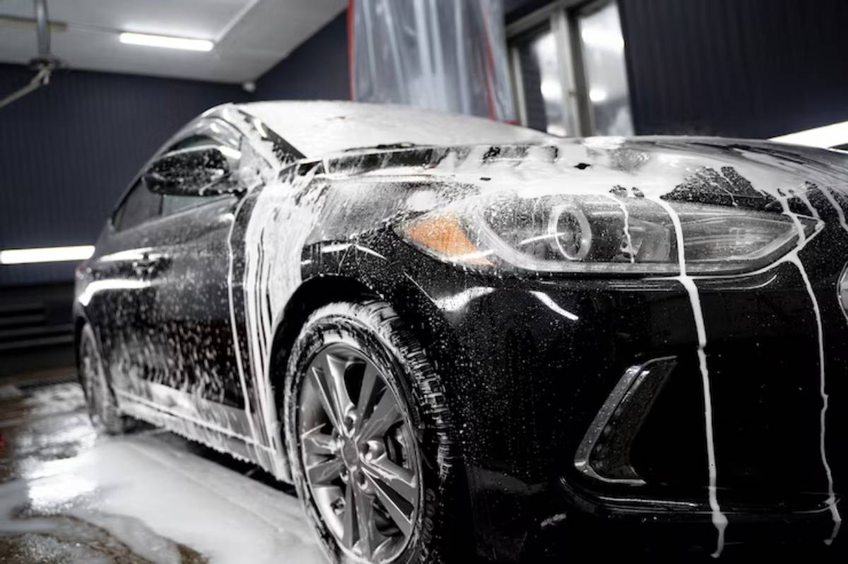 A black car covered in thick foam sits in a car wash, with soap suds dripping down its sides under bright indoor lighting.